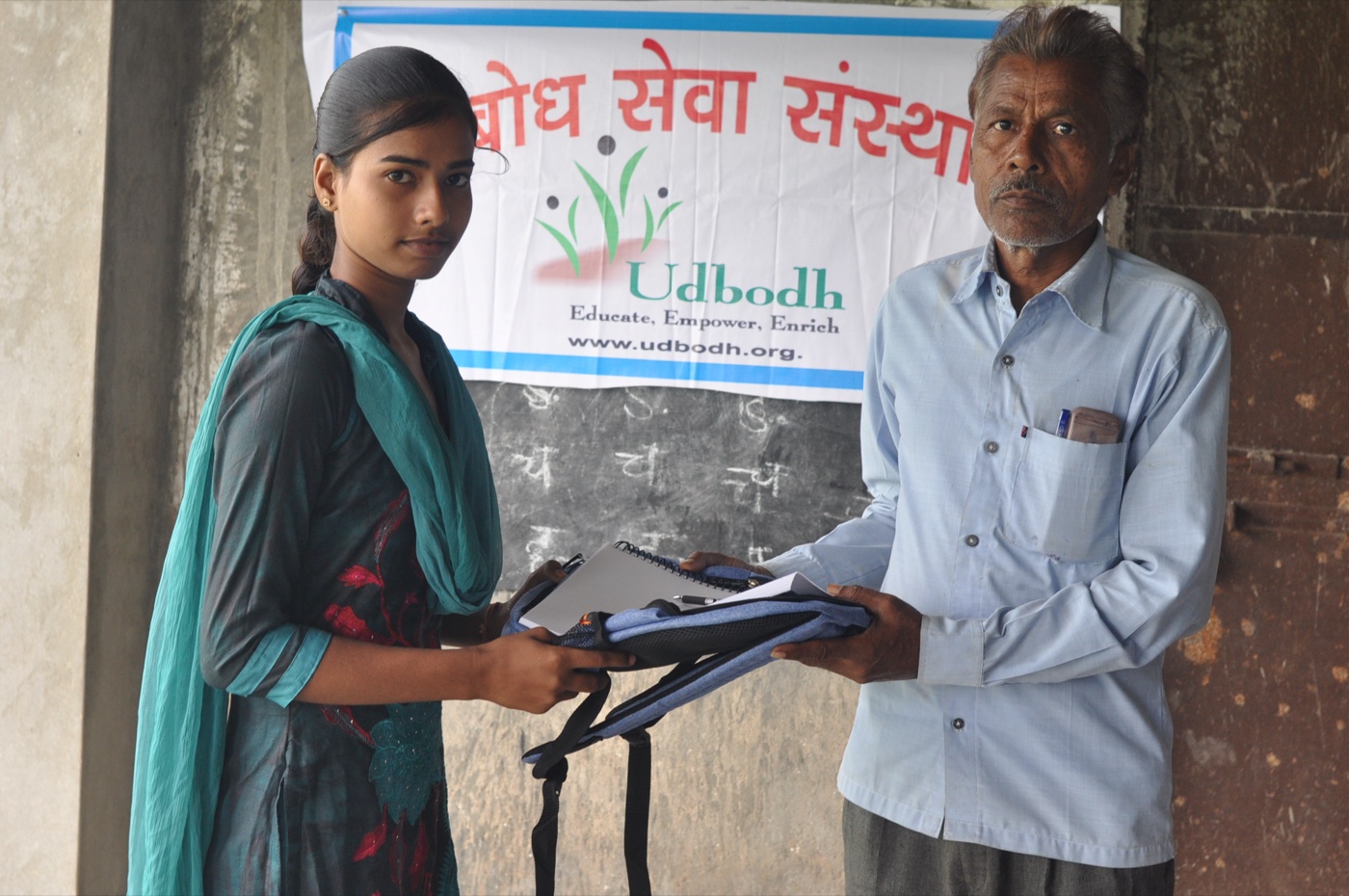 An Udbodh fellow handing a school bag and notebook to a community member in front of the Udbodh banner